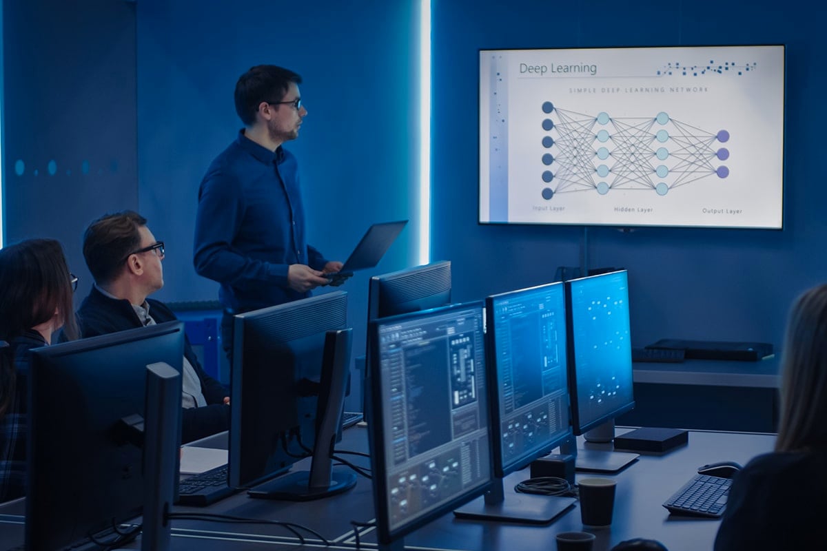 a man presenting on a large monitor to a room of individuals in front of their own computers