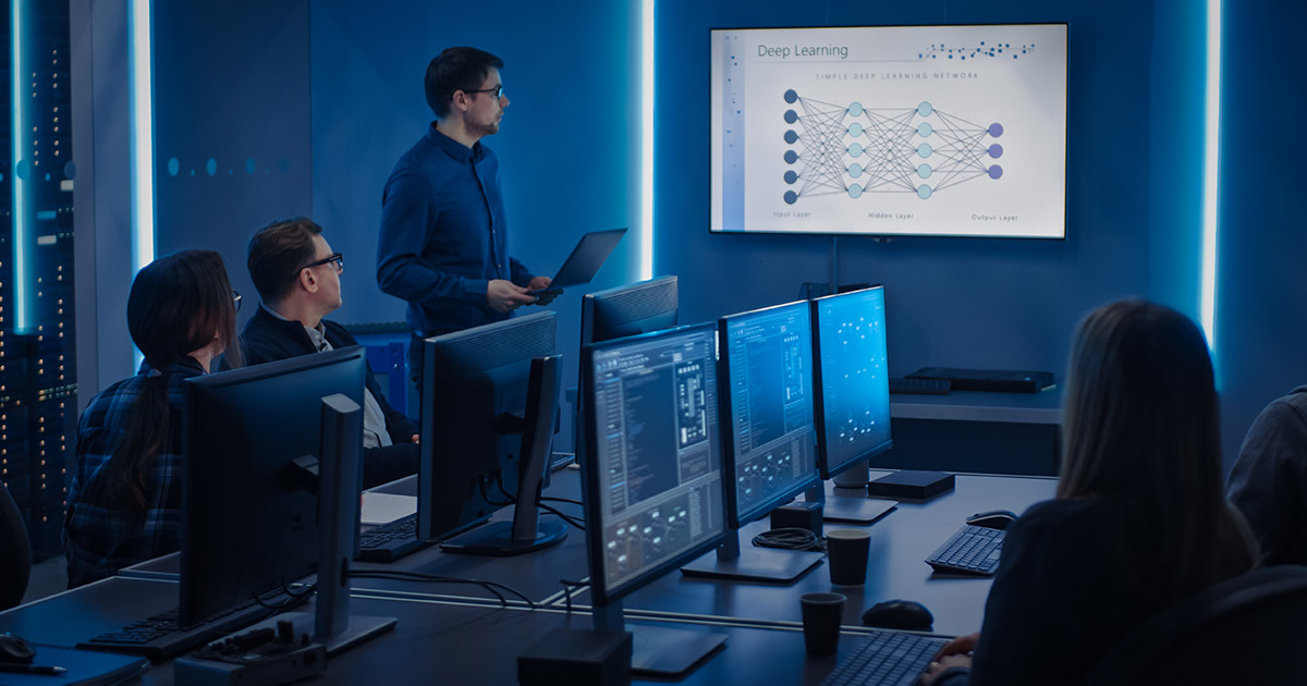 A group of people sitting in an office full of computers and monitors reviewing data on a large monitor.