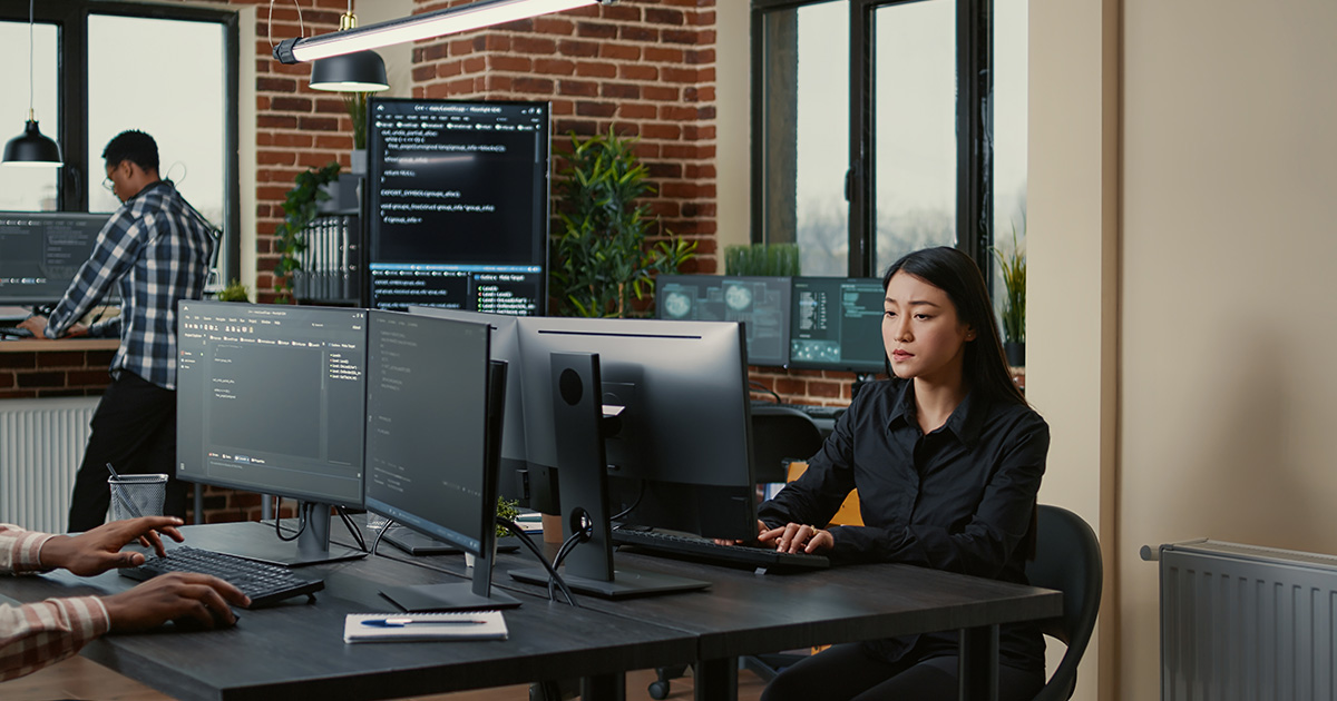 Cyber security expert with clipboard sitting down at desk in front of programer writing code while team of database admins compute big data