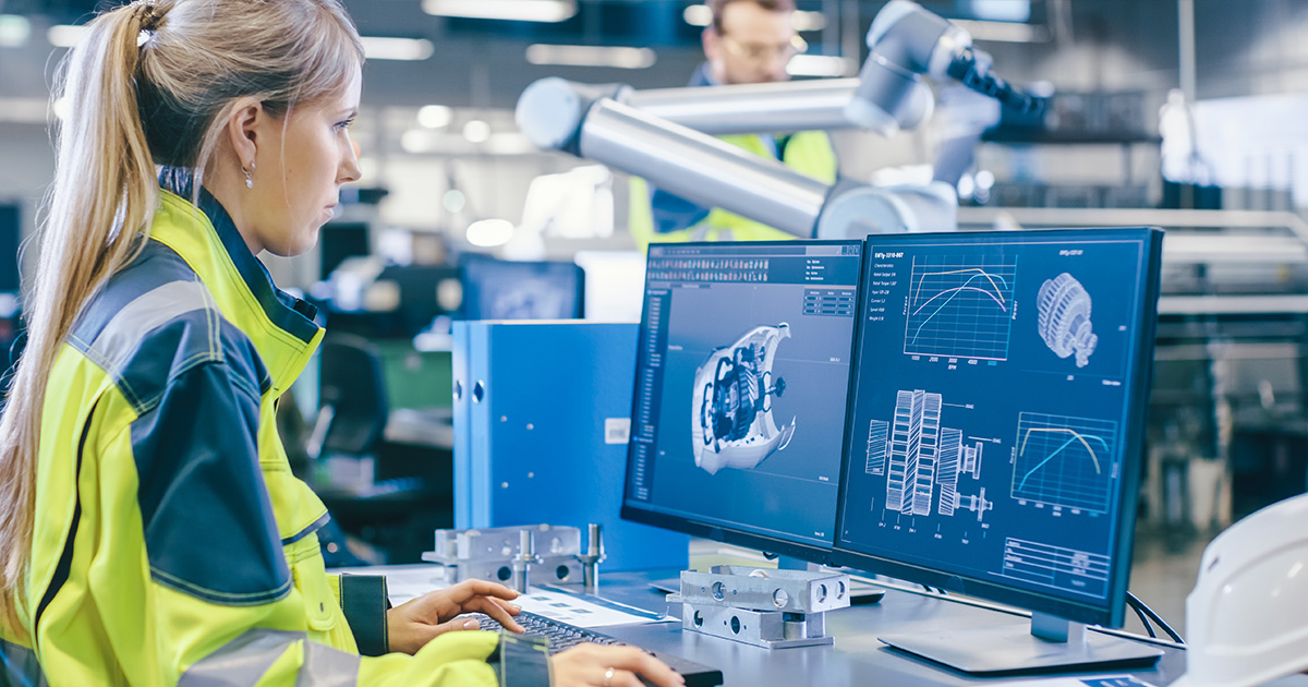 Female mechanical engineer in a yellow jacket works on her computer at a factory while a male automation engineer uses laptop for programming robotic arm in the background